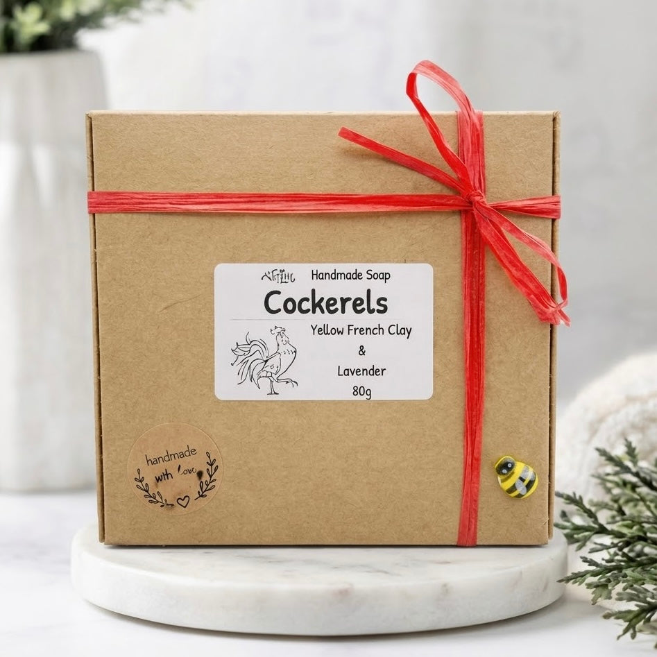 A square brown kraft gift box is tied neatly with red raffia ribbon into a simple bow. A white rectangle label is centered, reading "Handmade Soap, Cockerels," and lists "Yellow French Clay & Lavender, 80g" below, with a small bee charm attached to the corner.