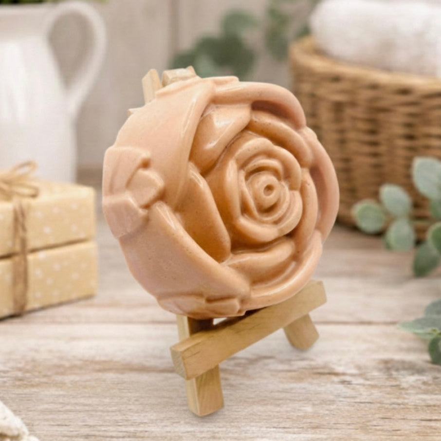 A handmade vegan soap, intricately sculpted in the shape of a large, blooming rose flower with layered petals, displayed on a small wooden easel against a bright win