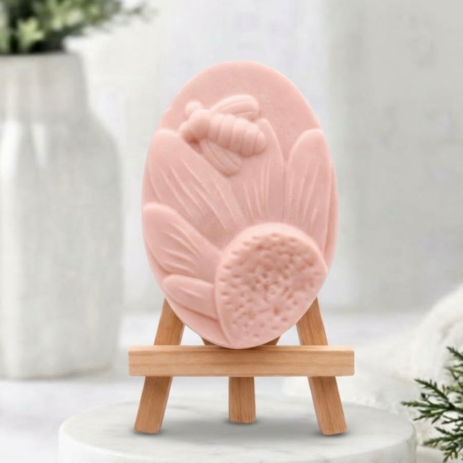 A soft pink soap bar shaped like a sunflower with a bee detail, displayed on a small wooden easel stand against a soft, blurred background.