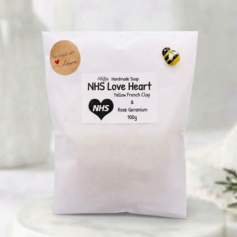 A packaged NHS Love Heart soap bar presented in a translucent white glassine paper bag. The package features a product label reading "NHS Love Heart - Yellow French Clay & Rose Geranium", a round kraft sticker stating "Handmade with love", and a decorative bumblebee detail on the corner.
