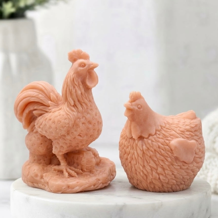 Two intricately detailed, terracotta coloured vegan soap bars - one cockerel and one hen - sitting on a white marble coaster with a bokeh background of a window and blurry greenery.