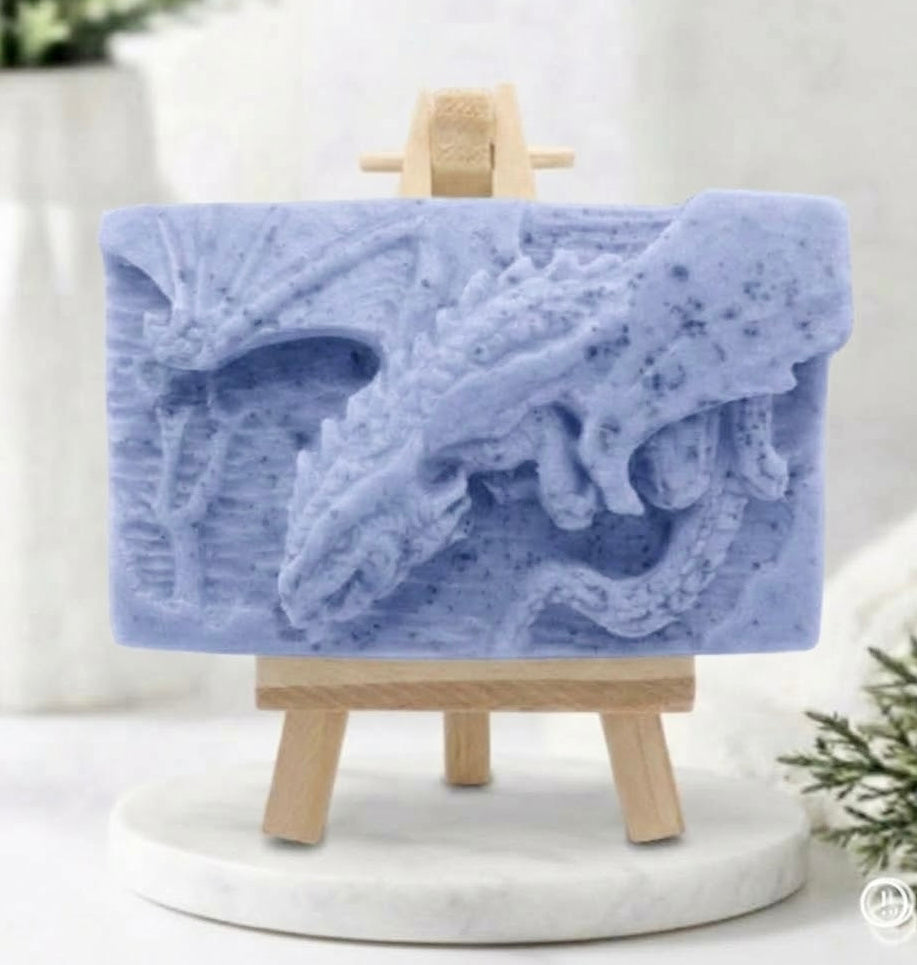 handmade soft blue soap bar featuring a detailed relief carving of a dragon, displayed on a small wooden easel in a bright, rustic room with white flowers in the background.