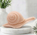A highly detailed handmade vegan soap in a soft pink colour, sculpted in the shape of a realistic snail with a prominent spiralled shell and a textured, leathery body, displayed on a light wooden surface.