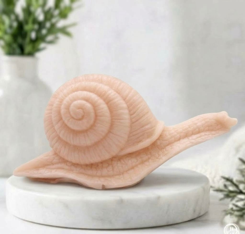 A highly detailed handmade vegan soap in a soft pink colour, sculpted in the shape of a realistic snail with a prominent spiralled shell and a textured, leathery body, displayed on a light wooden surface.