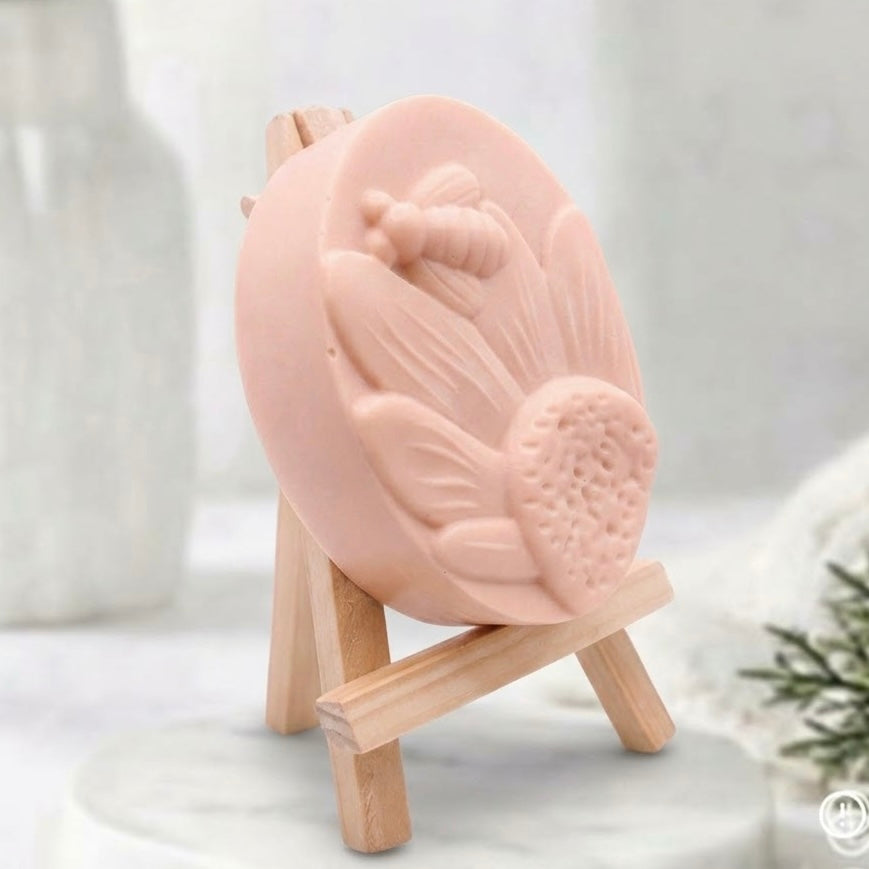 A pale pink oval soap bar with a raised bee and flower design, displayed on a small wooden easel stand against a softly blurred light background.