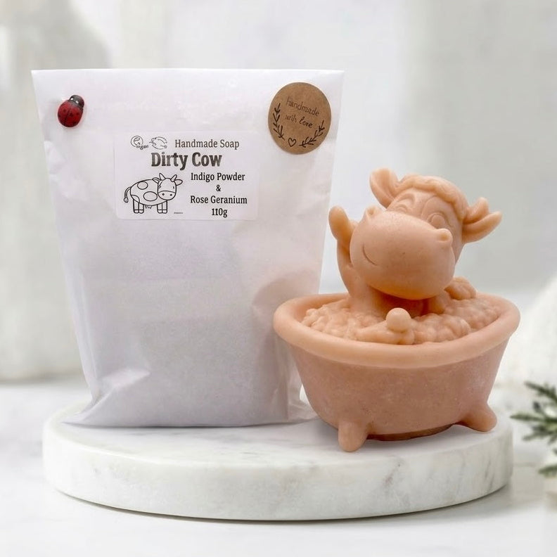 Novelty handmade vegan soap of a cute cow sitting in a bathtub, accompanied by its branded 'Dirty Cow' packaging bag with a ladybug pin and detailed ingredient list including Indigo Powder, Rose Geranium, and 110g weight.