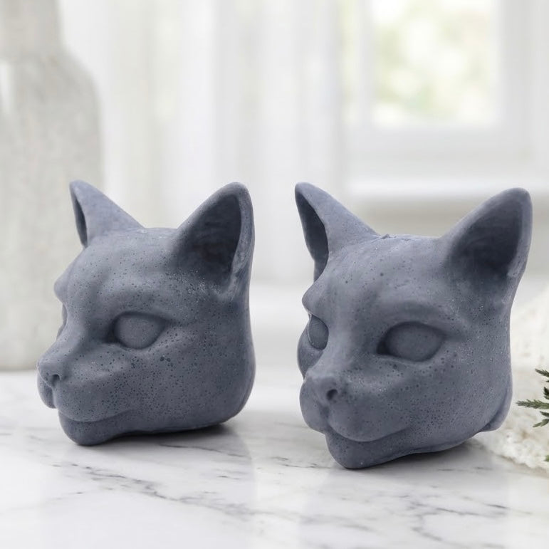Two charcoal grey, vegan soap bars handmade in the shape of Siamese cat heads, resting on white marble.