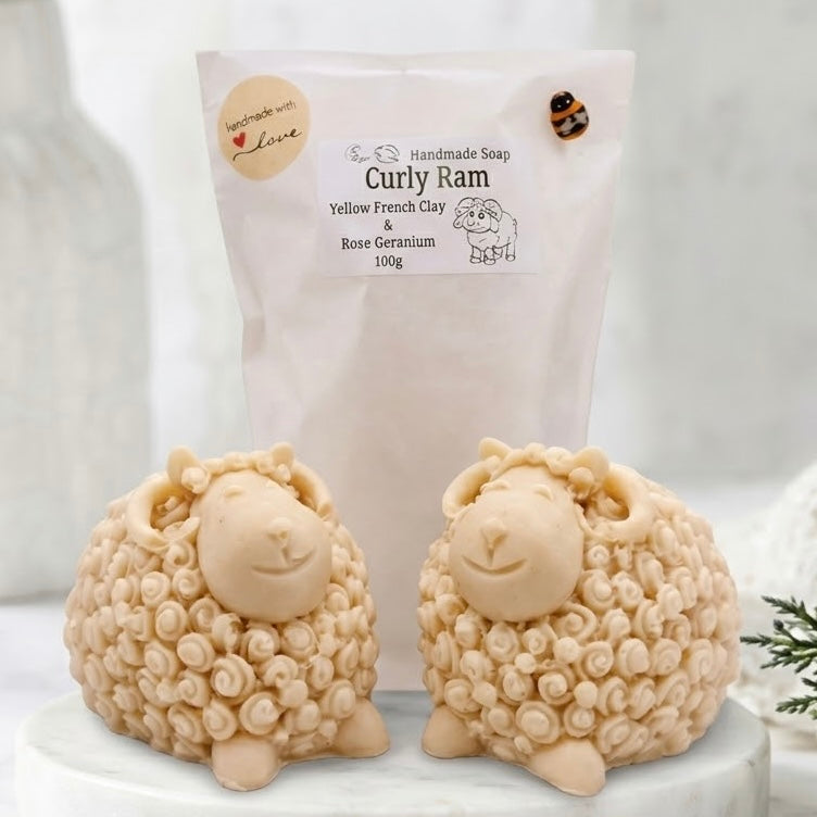 Two soft yellow sheep / Ram soaps with curly textured wool, closed eyes, and a smiling face, displayed on a white surface against a soft blurred background.