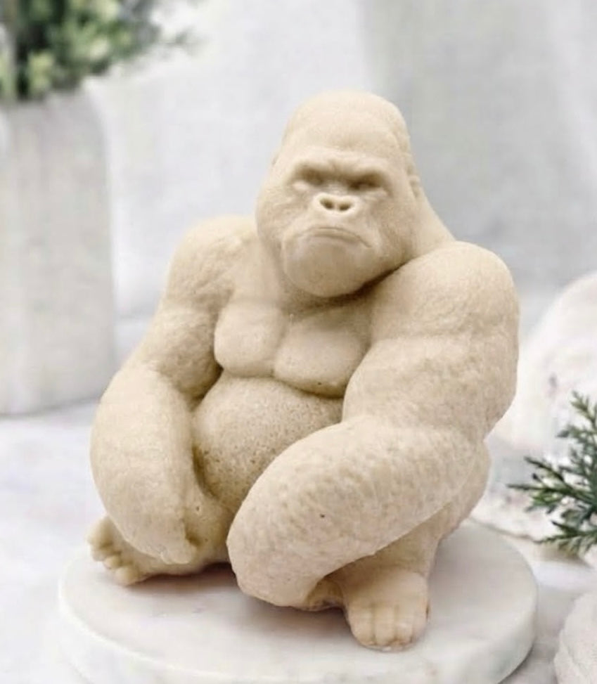 A yellow gorilla French clay vegan soap sitting on a round white marble base, photographed in a softly styled indoor setting with blurred plants and neutral decor in the background.