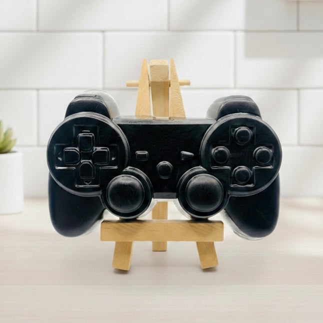 A 3D handmade vegan soap sculpted in the shape of a classic video game controller, finished in a deep charcoal black with realistic buttons and joysticks, displayed on a miniature wooden easel in a bright farmhouse setting.