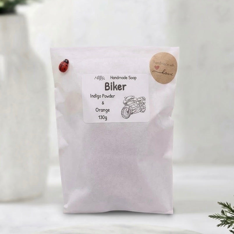 White paper packaging bag for 'Biker' handmade soap, containing Indigo Powder & Orange ingredients, featuring a motorcycle sketch and a red ladybug clip