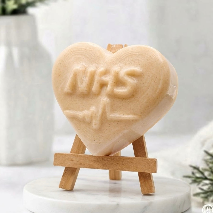 A side view of a soft yellow , heart-shaped soap featuring embossed "NHS" lettering and a heartbeat line below. The soap is displayed vertically on a small wooden easel set upon a round marble base. A white vase with greenery and evergreen sprigs rest in the softly blurred background