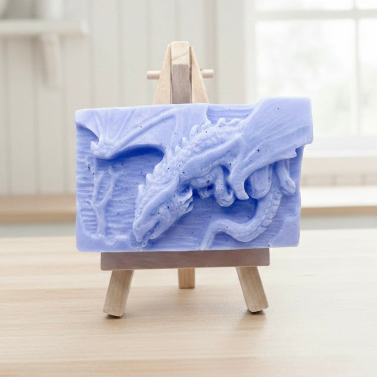 handmade soap bar featuring a detailed 3D relief carving of a dragon, displayed on a small wooden easel in a bright, rustic room with white flowers in the background.