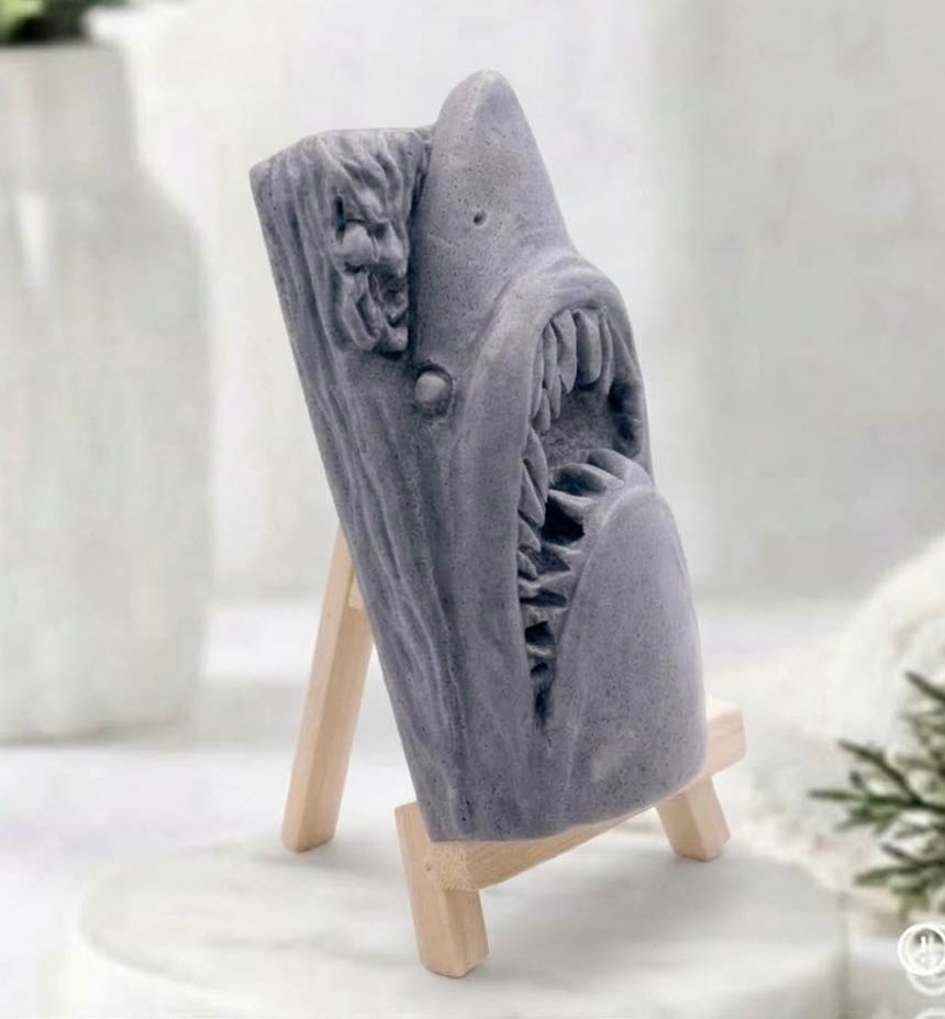 Side view of a handmade shark soap made with activated charcoal and displayed on a wooden easel against a neutral background featuring a white vase and greenery.