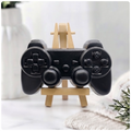 A handmade vegan soap sculpted in the shape of a classic video game controller, finished in a deep charcoal black with realistic buttons and joysticks, displayed on a miniature wooden easel in a bright farmhouse setting.