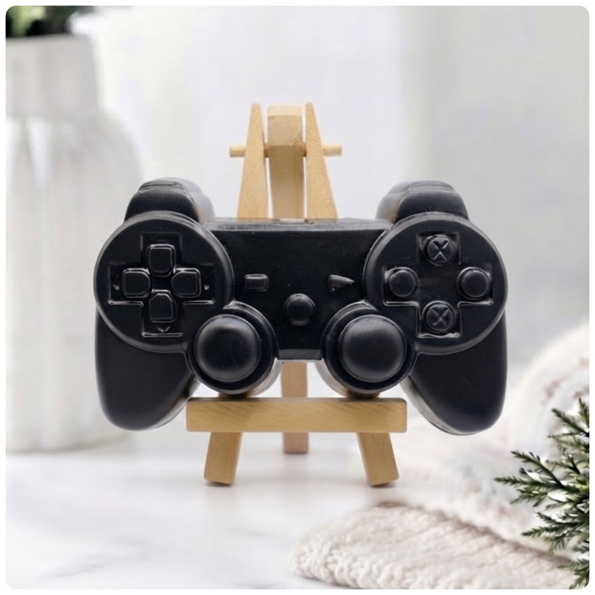 A handmade vegan soap sculpted in the shape of a classic video game controller, finished in a deep charcoal black with realistic buttons and joysticks, displayed on a miniature wooden easel in a bright farmhouse setting.