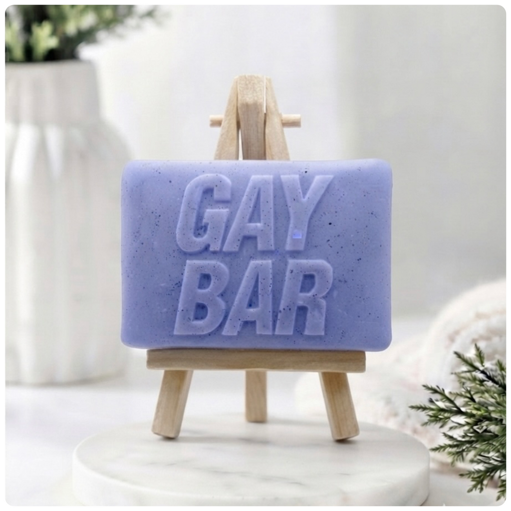 A rectangular handmade vegan soap featuring the words "GAY BAR" in large, bold, 3D block lettering, displayed on a miniature wooden easel.