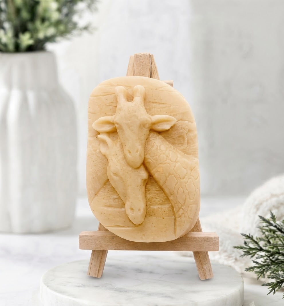 A close-up photograph of a light yellow-cream coloured handmade vegan soap sculpted into two detailed giraffe heads pressed together in a loving embrace. The soap is displayed on a small, light-wood artist's easel, resting on a white marble surface.