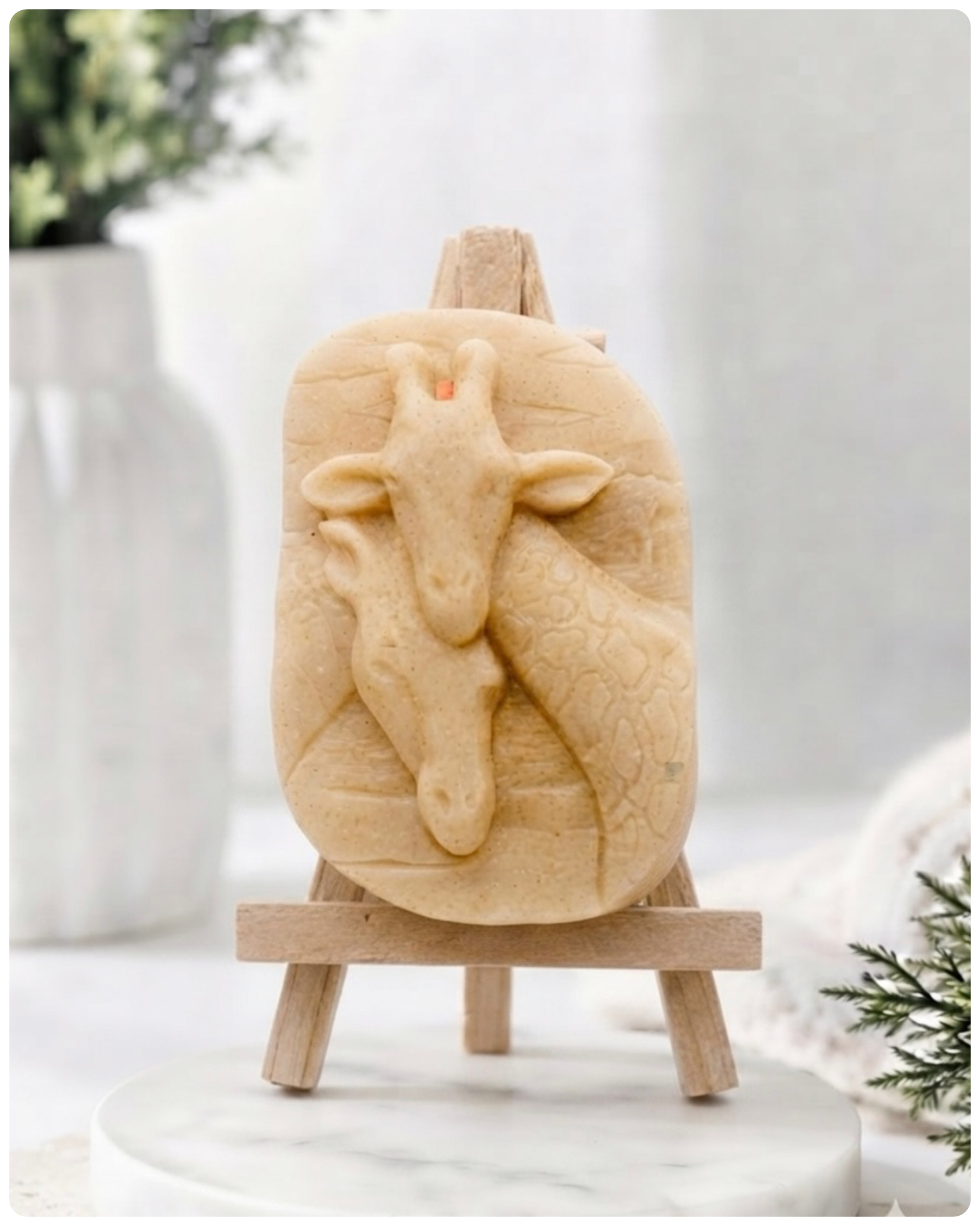 A rectangular handmade vegan soap in a natural cream tone, featuring a detailed relief of a mother and baby giraffe with their necks intertwined, displayed on a miniature wooden easel against a white wood-panelled background.
