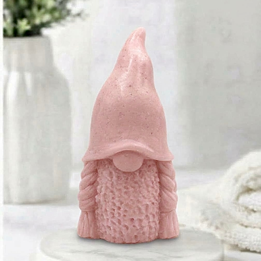 A handmade soft pink vegan soap in the shape of a female gnome (or gonk), featuring a tall, slightly curved hat pulled over the eyes and two thick, textured plaits framing a textured dress.