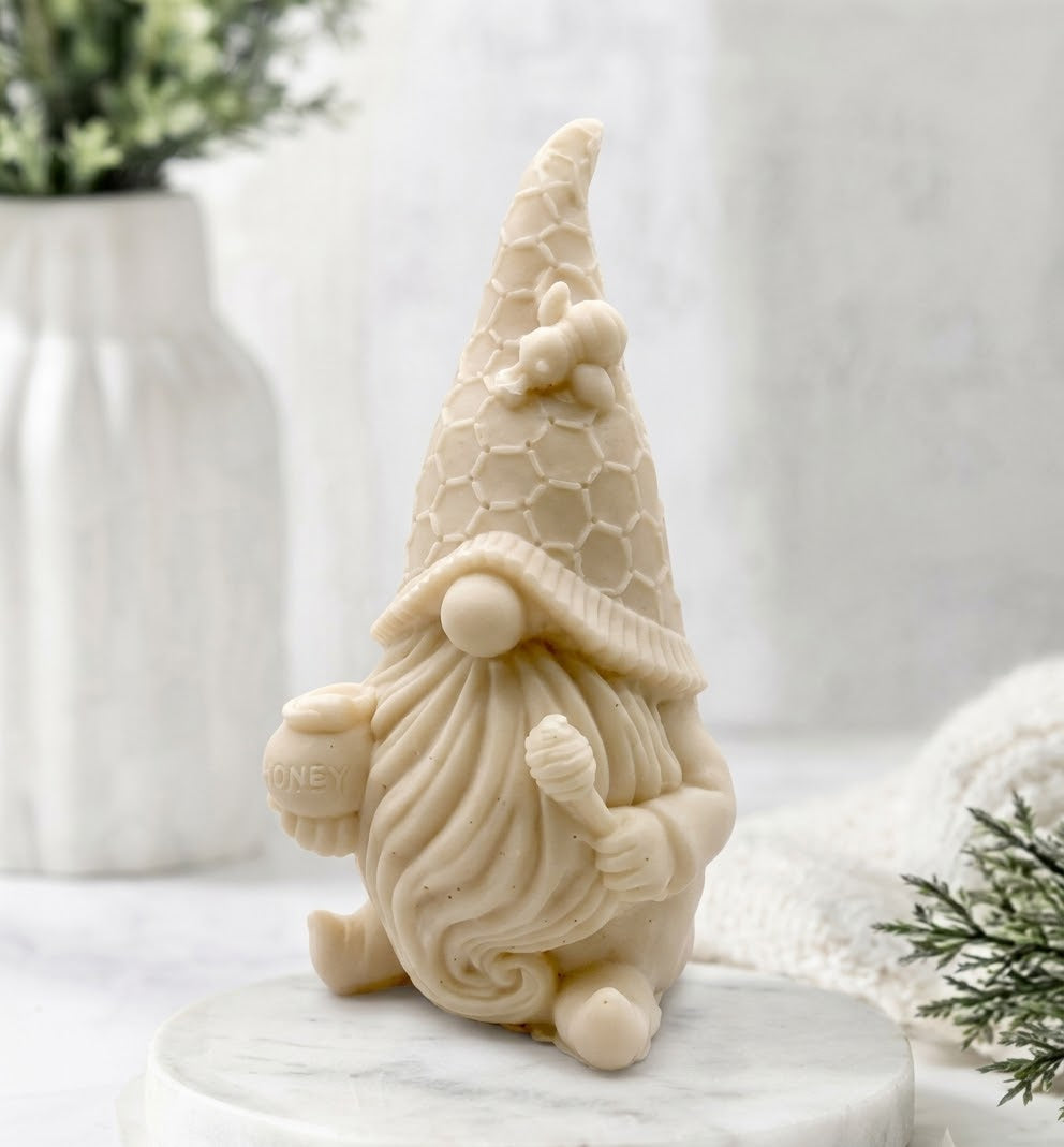 A close-up photograph of a detailed cream-coloured gnome-shaped vegan soap with a honeycomb-patterned hat and a small bee, resting on a white marble coaster in a brightly lit bathroom setting.