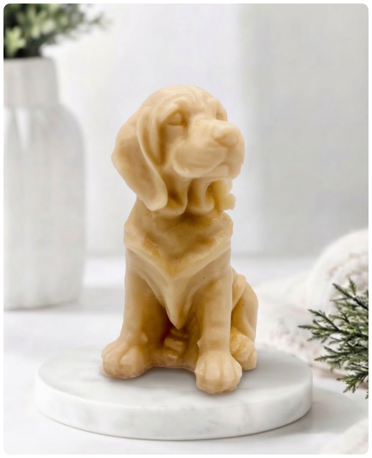 A yellow golden retriever dog-shaped soap sitting on a round white marble base, photographed in a softly styled indoor setting with blurred white vases and greenery in the background.