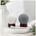 A handmade vegan golf ball soap gift set featuring one white and one grey spherical soap with realistic dimpled textures, displayed on small wooden stands next to a white golf tee and a kraft gift box tied with a red ribbon