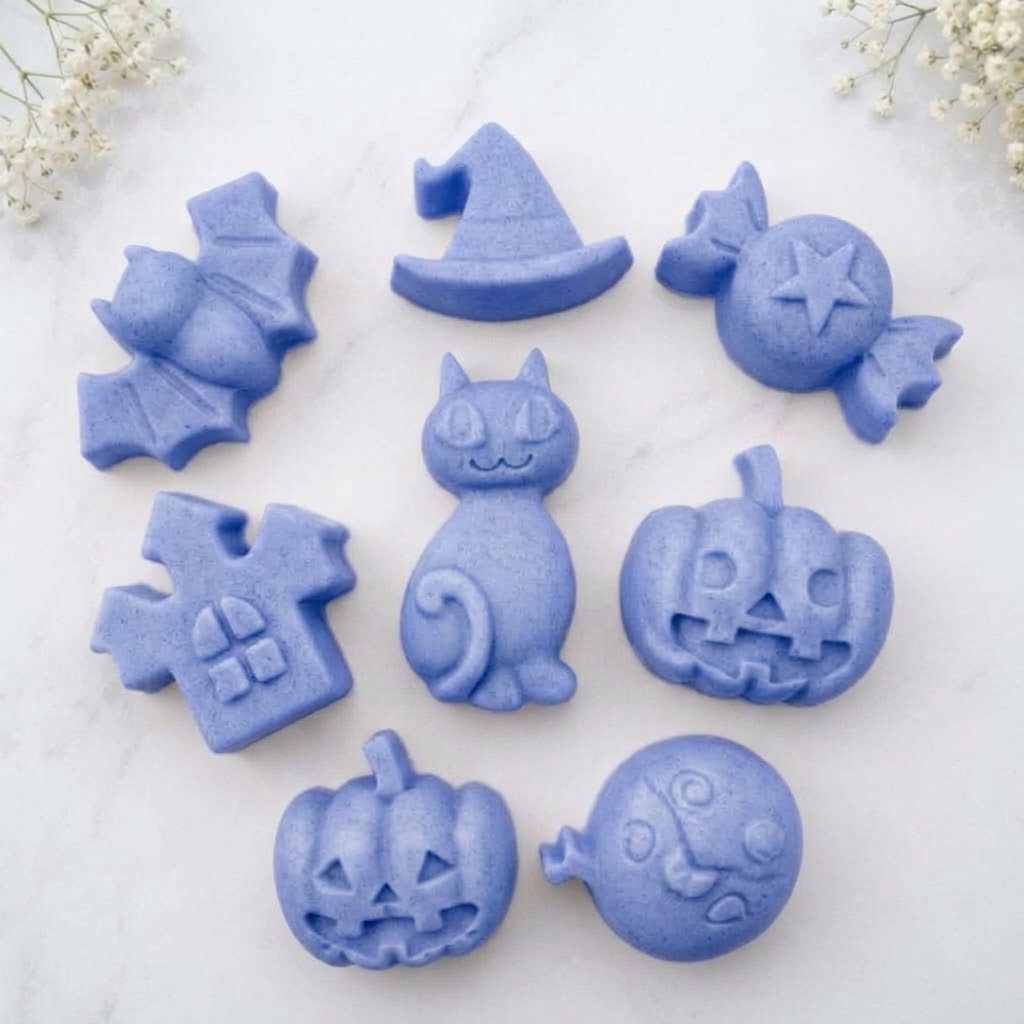 A soft blue collection of eight handmade vegan Halloween soaps featuring a bat, witch's hat, star-patterned sweet, haunted house, black cat, jack-o'-lantern, and a ghost, arranged on a light wooden surface with white floral accents.