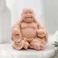 A handmade vegan soap sculpted into a joyful Laughing Buddha sitting in a meditative pose, holding prayer beads and a small bowl, displayed in a bright farmhouse setting