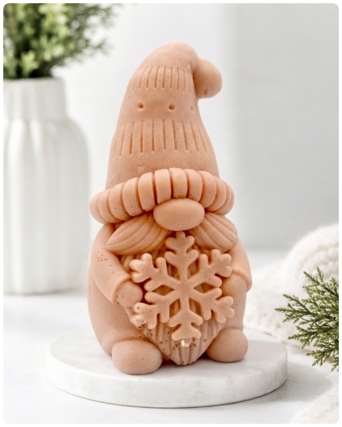 handmade vegan soap in the shape of a winter gnome sculpture holding a large snowflake, featuring a tall textured hat and a bushy moustache , displayed on a rustic white wooden surface with daisies and a milk churn in the background.