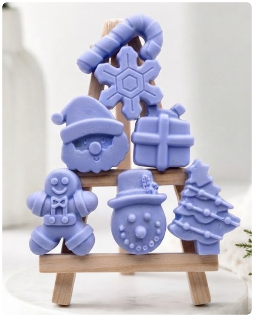 A collection of six handmade vegan Christmas soaps featuring a candy cane, snowflake, Santa Claus face, wrapped gift box, gingerbread man, snowman, and a decorated Christmas tree, arranged in a pyramid on a wooden stand.