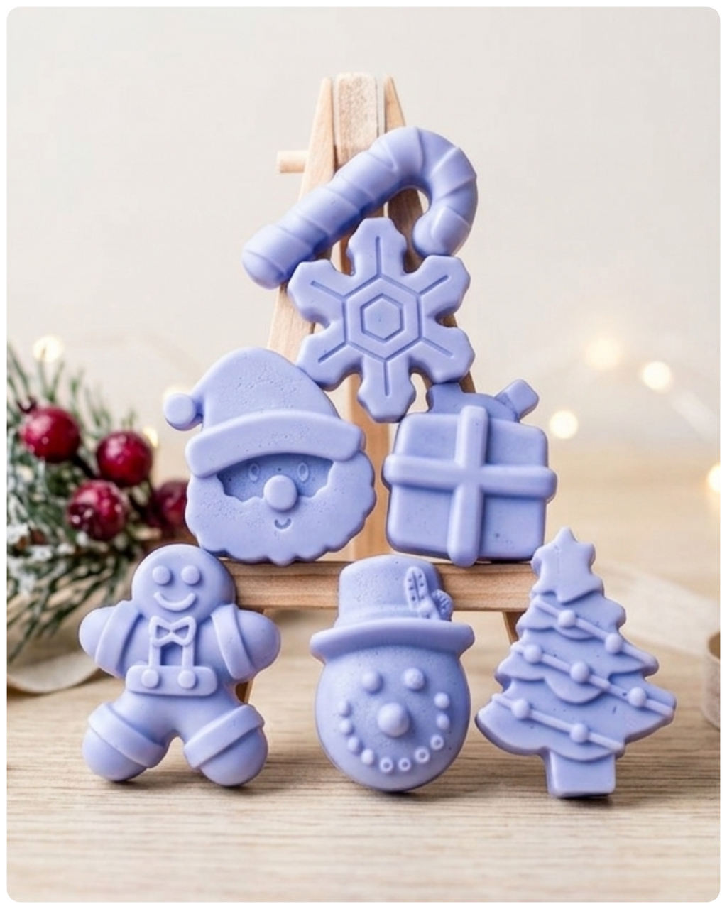 A collection of six handmade vegan Christmas soaps featuring a candy cane, snowflake, Santa Claus face, wrapped gift box, gingerbread man, snowman, and a decorated Christmas tree, arranged in a pyramid on a wooden stand.