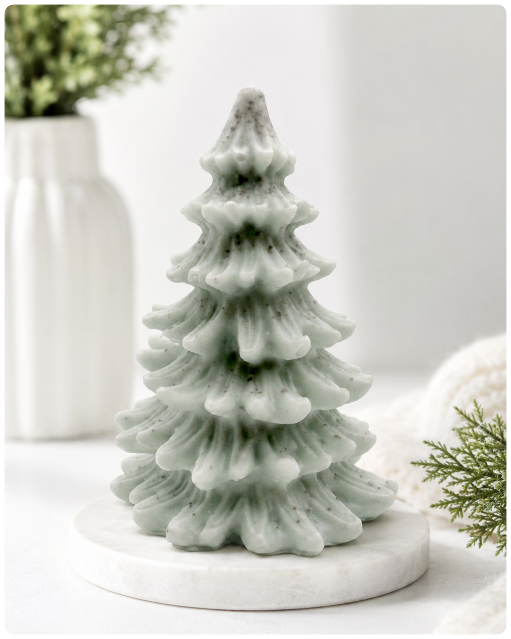 A tiered Christmas tree soap in a pale sage green colour with dark speckles, sitting on a round white marble base. The background is a soft, bright white with blurred decorative props including a white vase with greenery on the left and a white knit blanket with pine branches on the right.