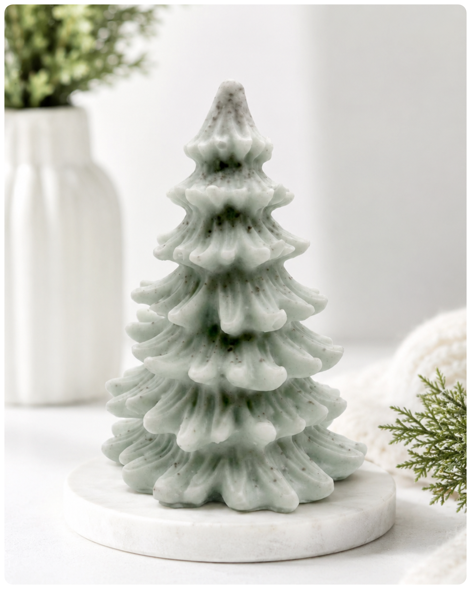 A tiered Christmas tree soap in a pale sage green colour with dark speckles, sitting on a round white marble base. The background is a soft, bright white with blurred decorative props including a white vase with greenery on the left and a white knit blanket with pine branches on the right.