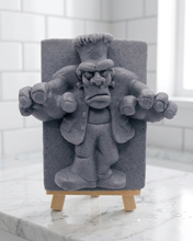 Load image into Gallery viewer, A rectangular handmade vegan soap featuring a high-relief carving of Frankenstein's monster in a classic pose with arms outstretched and a grumpy expression, displayed on a miniature wooden easel.
