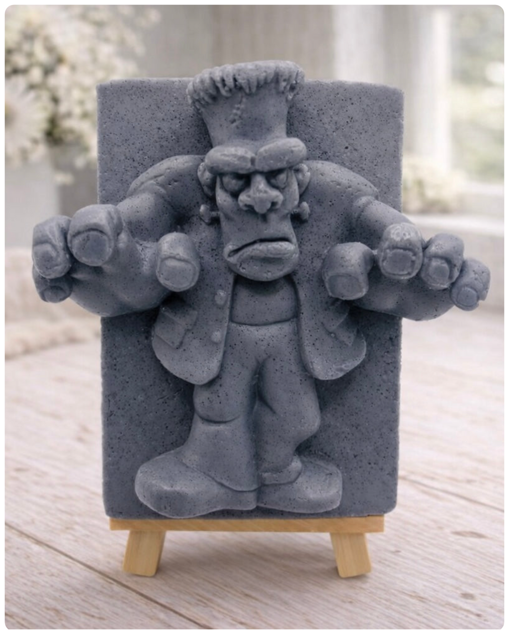 A rectangular handmade vegan soap featuring a high-relief carving of Frankenstein's monster in a classic pose with arms outstretched and a grumpy expression, displayed on a miniature wooden easel.