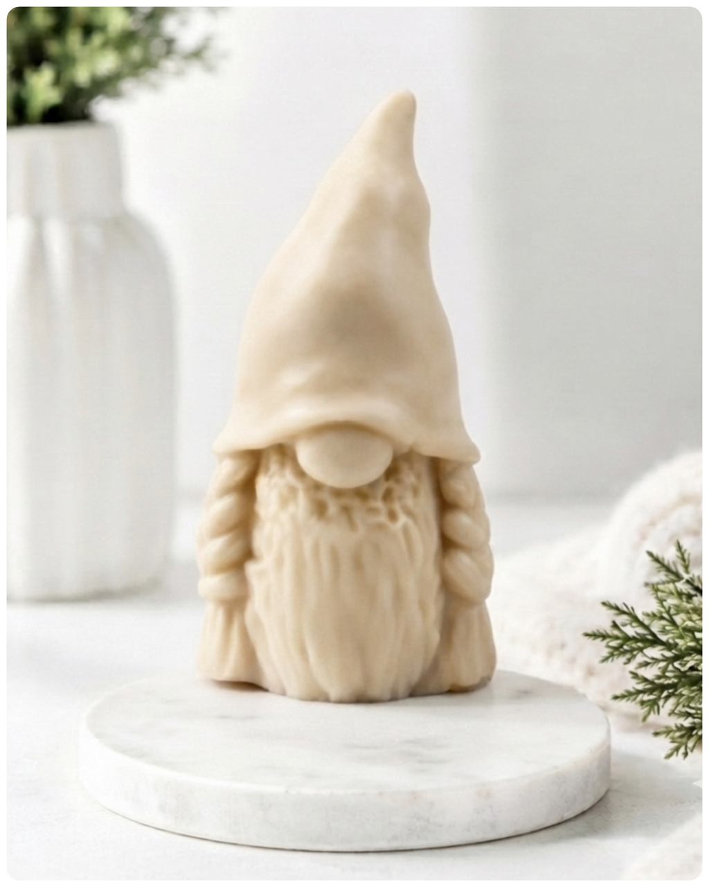 A handmade vegan soap sculpted in the shape of a female gnome (or gonk), featuring a tall, slightly curved hat pulled over the eyes and two thick, textured plaits framing a textured dress, in a natural cream colour.