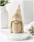 A handmade vegan soap sculpted in the shape of a female gnome (or gonk), featuring a tall, slightly curved hat pulled over the eyes and two thick, textured plaits framing a textured dress, in a natural cream colour.