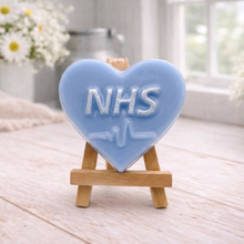 Load image into Gallery viewer, handmade vegan soap in a heart shape featuring an "NHS" relief and a heartbeat pulse line, displayed on a small wooden easel in a bright farmhouse decor setting.
