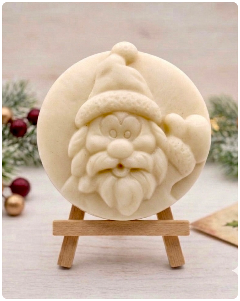 A circular handmade vegan soap featuring a detailed high-relief carving of a jolly Father Christmas waving, with a textured beard and a classic Santa hat, displayed on a miniature wooden easel.