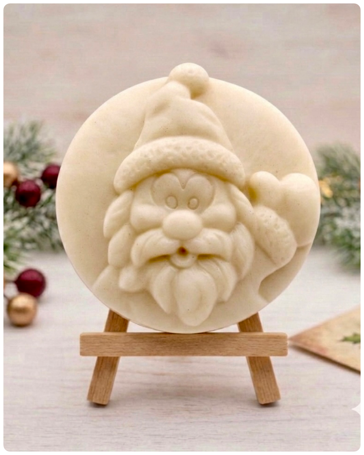 A circular handmade vegan soap featuring a detailed high-relief carving of a jolly Father Christmas waving, with a textured beard and a classic Santa hat, displayed on a miniature wooden easel.