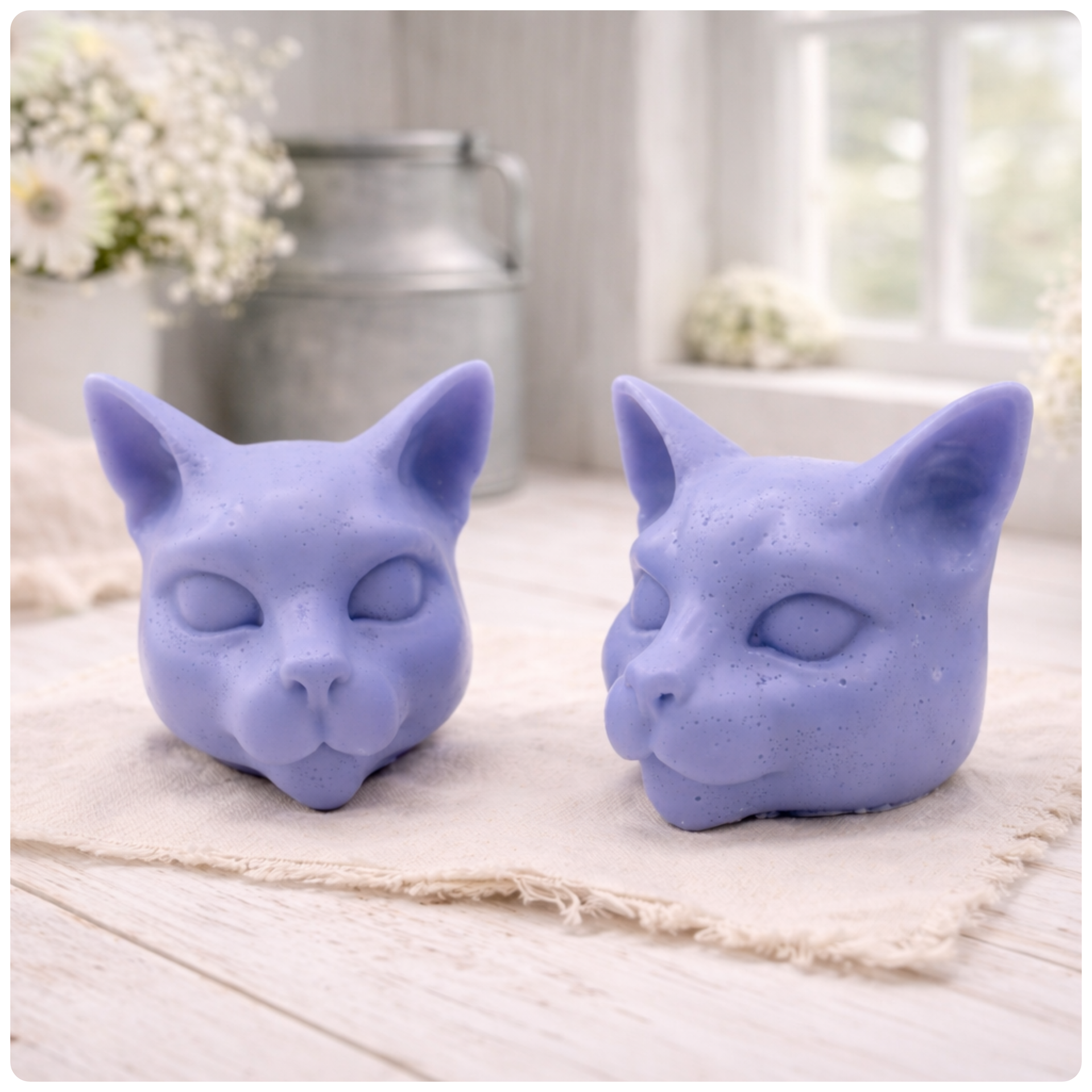 two handmade vegan soaps in the shape of Siamese cat heads, featuring elegant pointed ears and sleek facial features, displayed on a neutral cloth in a bright farmhouse decor setting.