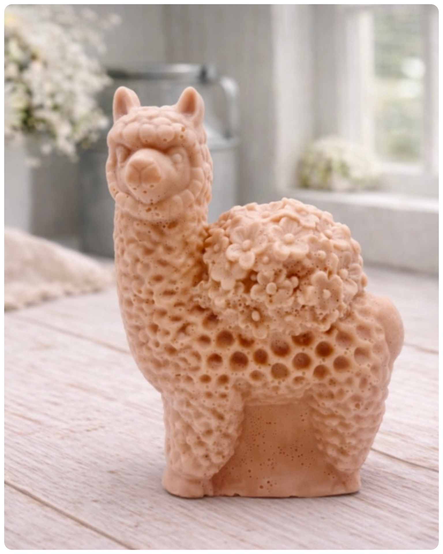 A handmade vegan soap featuring a charming llama or alpaca with a highly textured, fleecy coat and a blanket of intricate flowers across its back, displayed on a light wooden surface in a bright room.