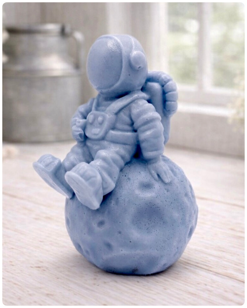 handmade vegan soap in the shape of a realistic astronaut sculpture sitting on the moon, featuring detailed spacesuit textures and craters on the lunar surface, displayed in a bright farmhouse decor setting.