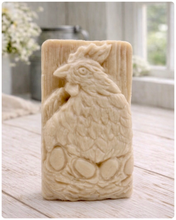 Load image into Gallery viewer, A rectangular handmade vegan soap in a natural colour, featuring a highly detailed relief carving of a broody hen chicken sitting on a nest of eggs, displayed upright on a light wooden surface.
