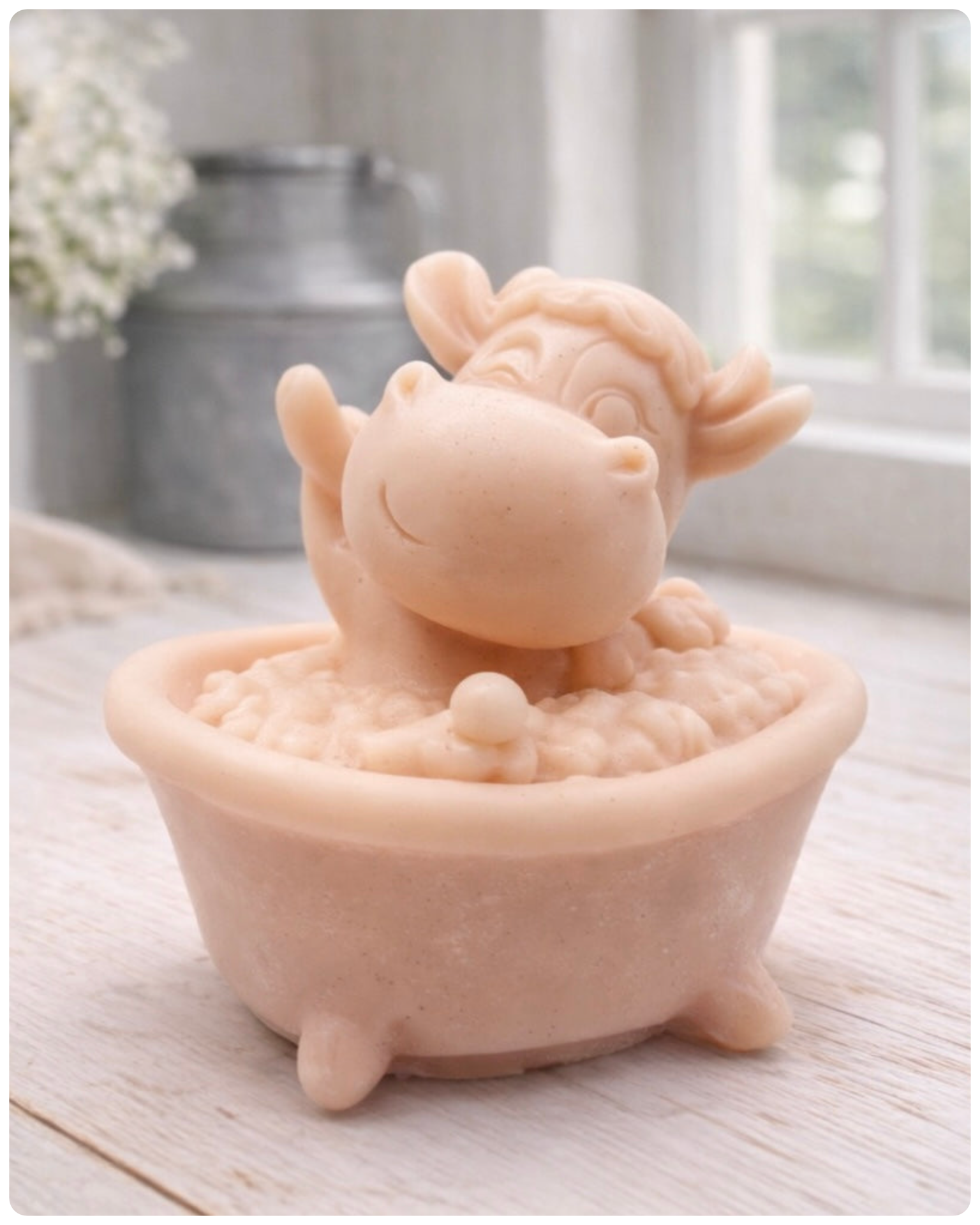 A handmade vegan soap is sculpted in the shape of a "dirty cow" character happily sitting in a bubble-filled roll-top bathtub with one hoof raised, displayed on a light wooden surface.