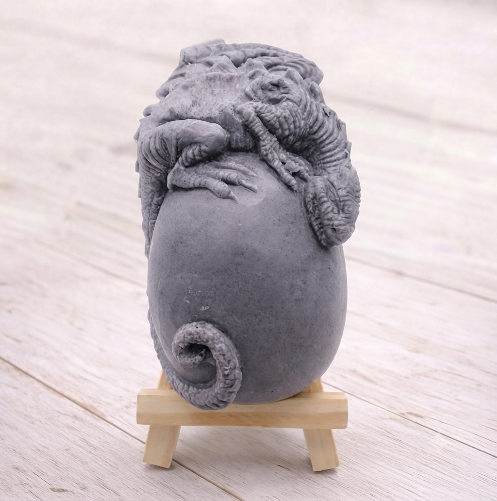 A 3D handmade vegan soap in a charcoal-grey tone, sculpted as a large prehistoric egg with a small dinosaur or dragon clinging to the top, featuring detailed scales and a curled tail, displayed on a miniature wooden easel.