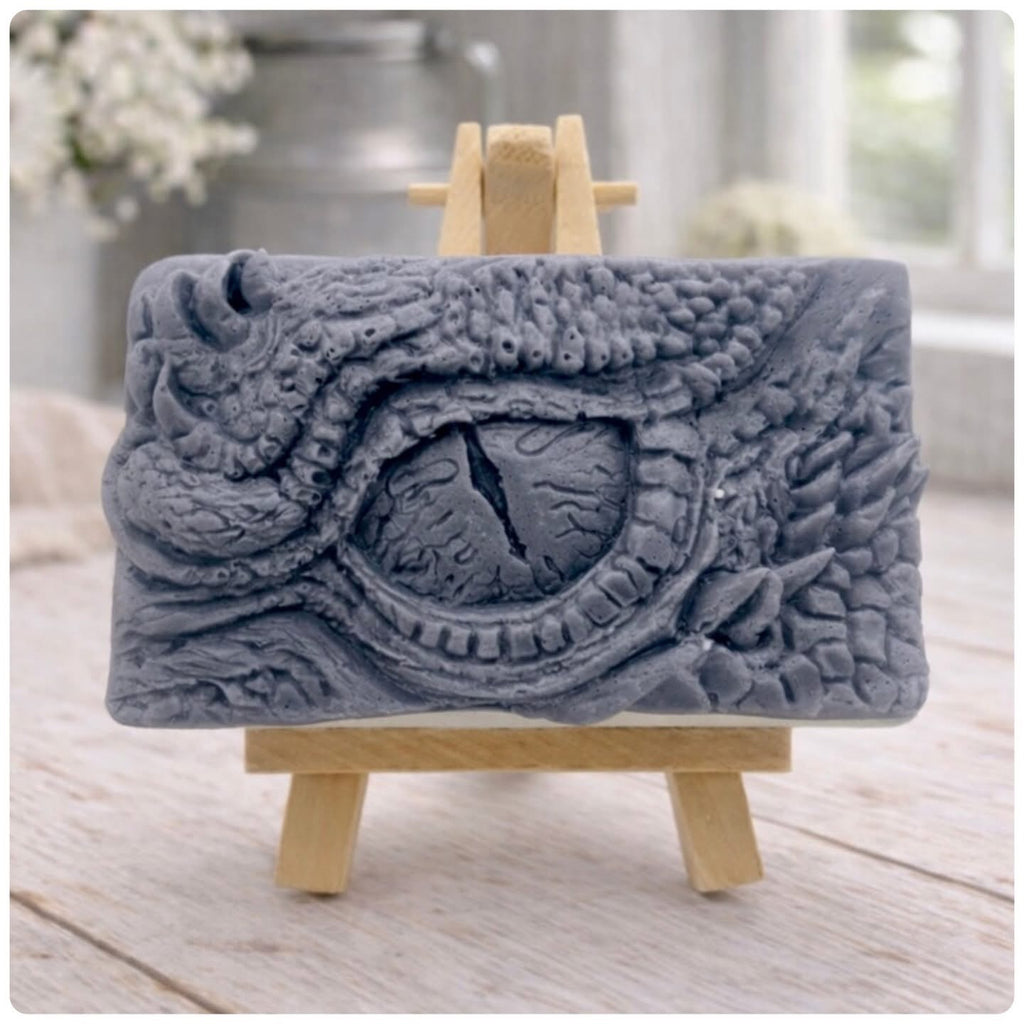A rectangular handmade vegan soap in a slate-grey tone, featuring a highly detailed, close-up relief of a mythical dragon or dinosaur eye surrounded by thick, pebbled scales and reptilian skin textures, displayed on a miniature wooden easel.