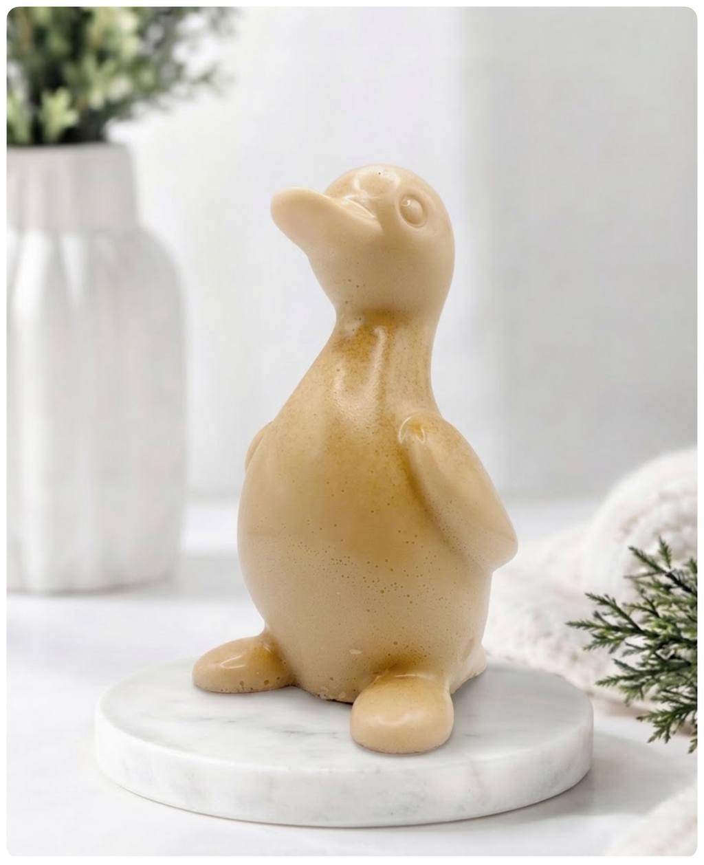 A chunky yellow duck vegan soap with a speckled finish, sitting on a round white marble base. The background is a softly blurred, bright interior with a white vase and greenery.
