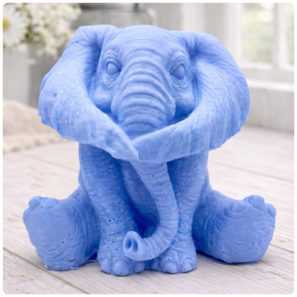 A highly detailed handmade vegan soap in a charcoal grey tone, sculpted in the shape of a sitting elephant with realistic wrinkled skin textures and large ears, displayed on a light wooden surface with white flowers in the background.